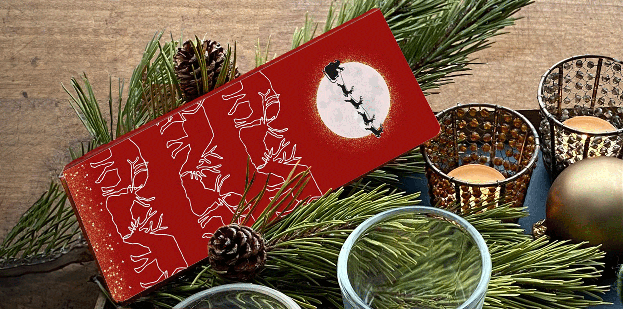 Rote CHOCQLATE Weihnachtsschatulle mit Rentieren und Santa Claus vor dem Mond auf Kiefernzweigen, daneben Windlichter und eine goldene Kugel