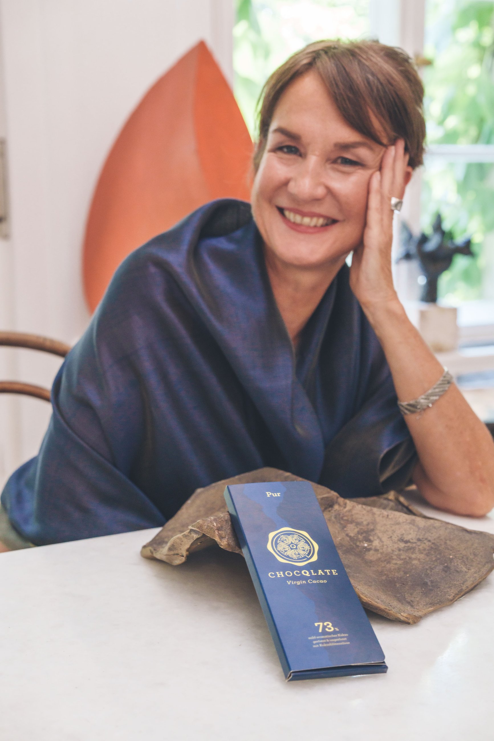 Porträt von Julia: lachend, mit kurzem dunkelbraunem Haar und dunkelblauem Seidentuch, die Wange in die Hand gestützt, vor ihr eine Tafel CHOCQLATE „Pur“ 73 % an einem Stück Kakaomasse
