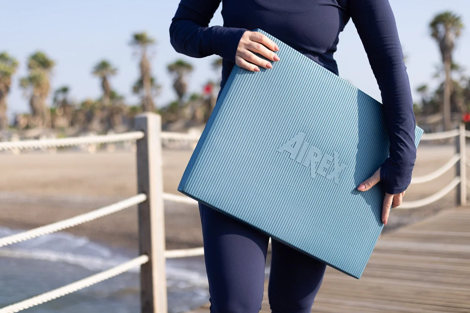 Frau in dunkelblauer Sportkleidung trägt ein blaues AIREX Balance-pad auf einem Holzsteg am Strand