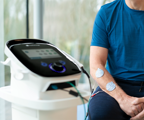 Appareil multifonction Chattanooga avec écran tactile monté sur un chariot, à côté duquel se trouve un patient muni d'électrodes sur l'avant-bras