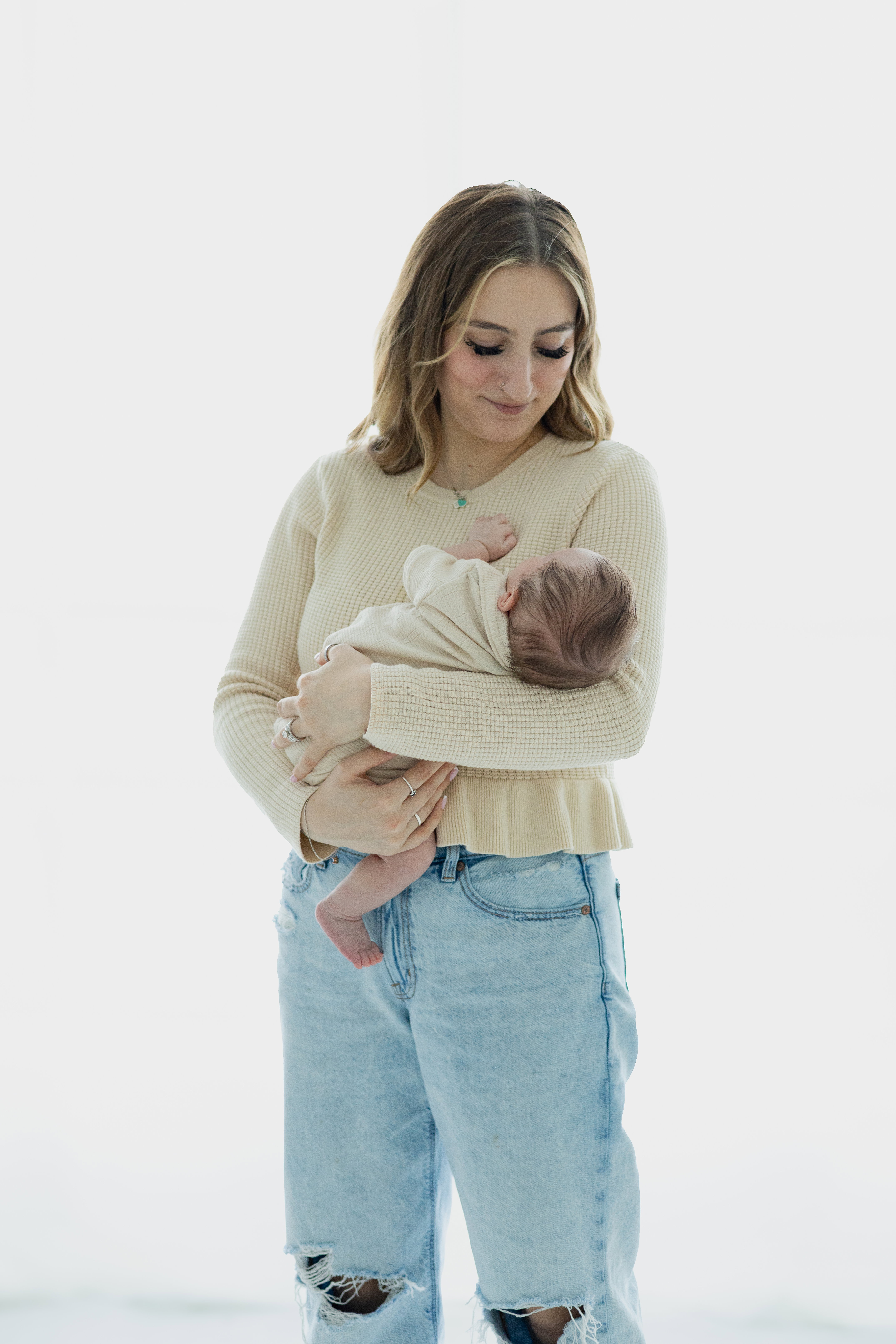 A mother in a cream sweater working at an embroidery machine in a sunlit home studio while her toddler plays with thread spools on the floor beside her