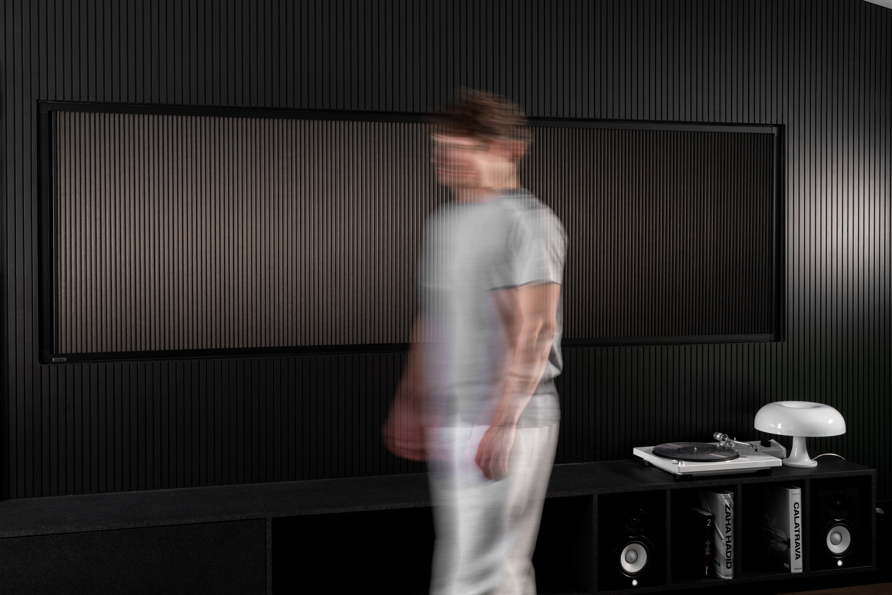 Man walking past a dark slatted wall with a record player and speakers on a low black cabinet