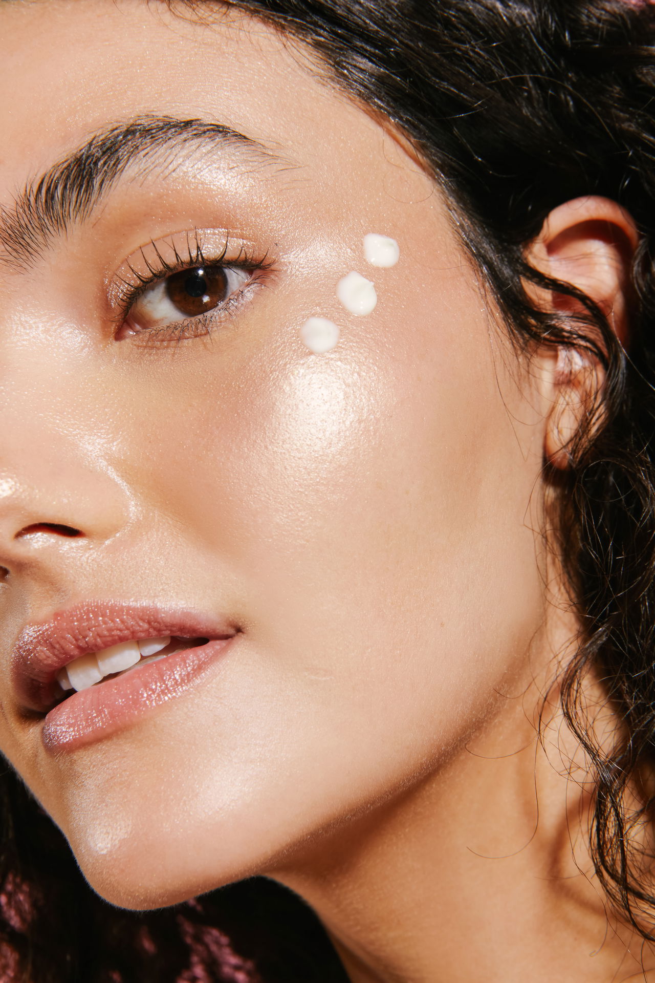 Close-up of a woman's face with three dots of cream near the eye on smooth, glowing skin.