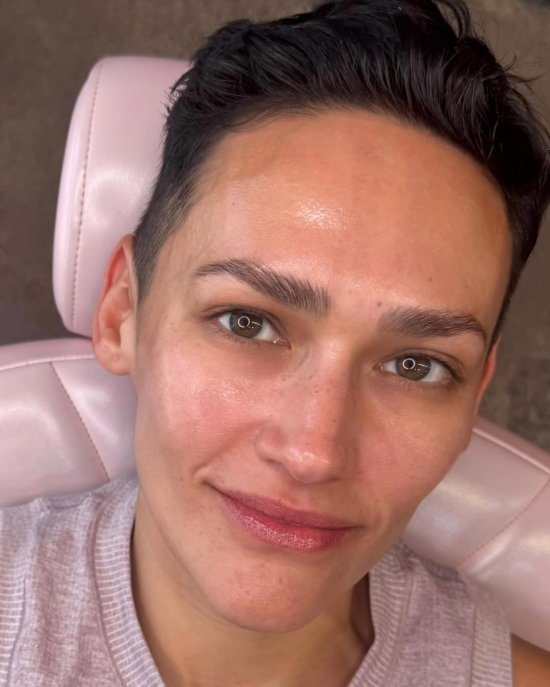 Person with short hair and natural makeup reclining on a pink chair, wearing a light sweater.