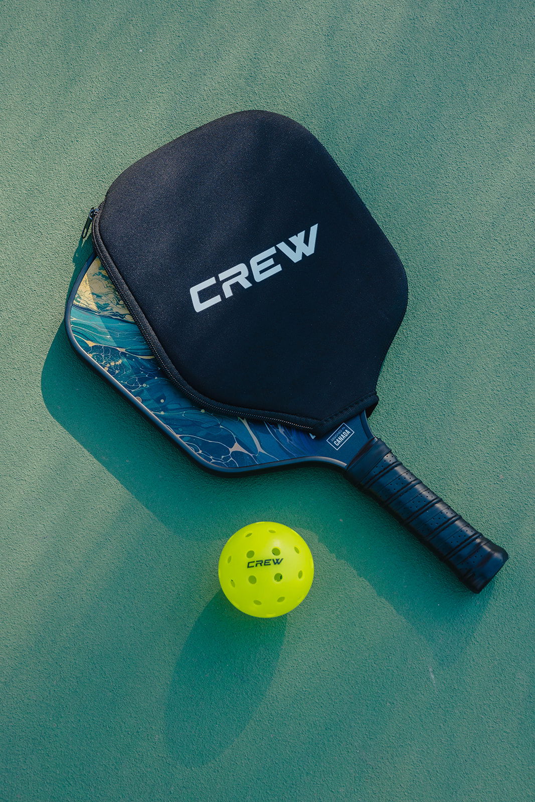 Black pickleball paddle with &quot;CREW&quot; cover and a neon yellow ball on green court surface.