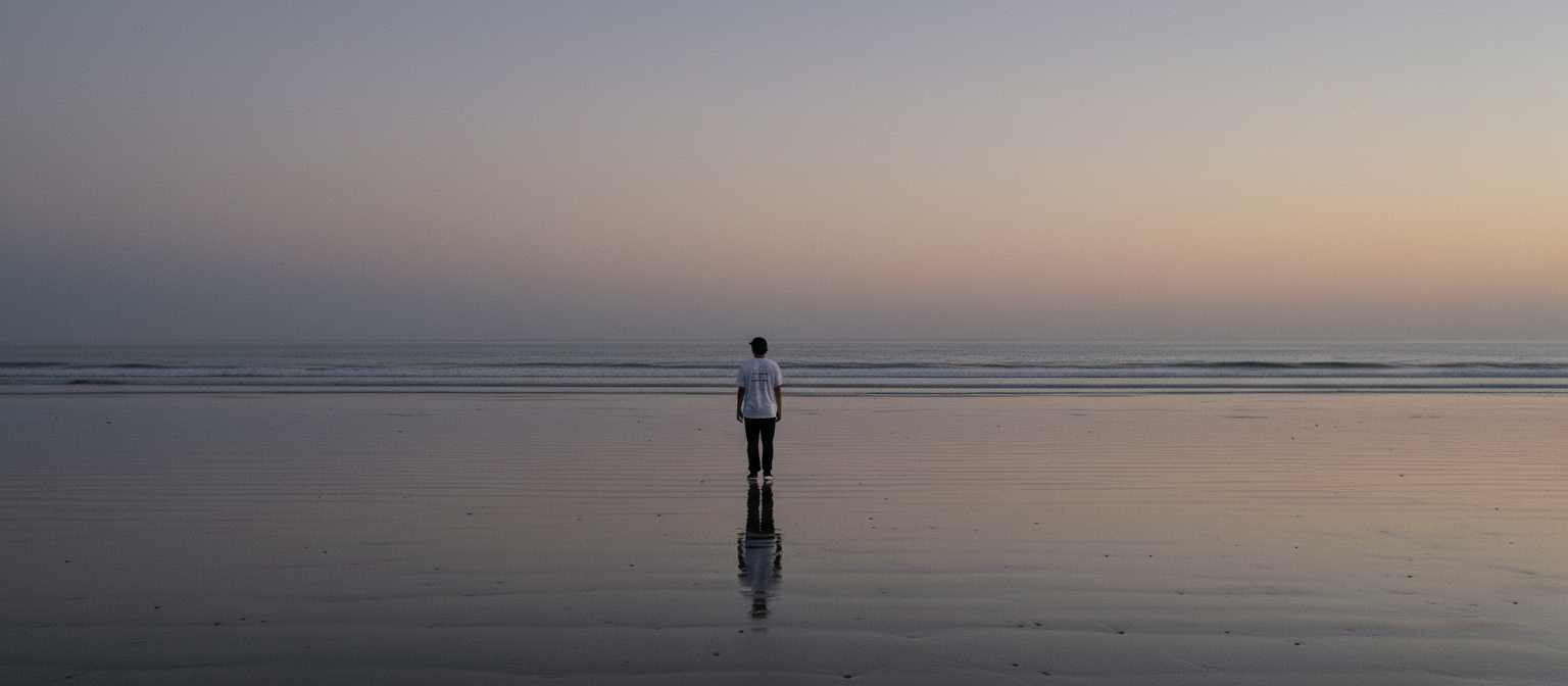 A lone figure in a white tee at the ocean's edge at dusk, reflected on wet sand