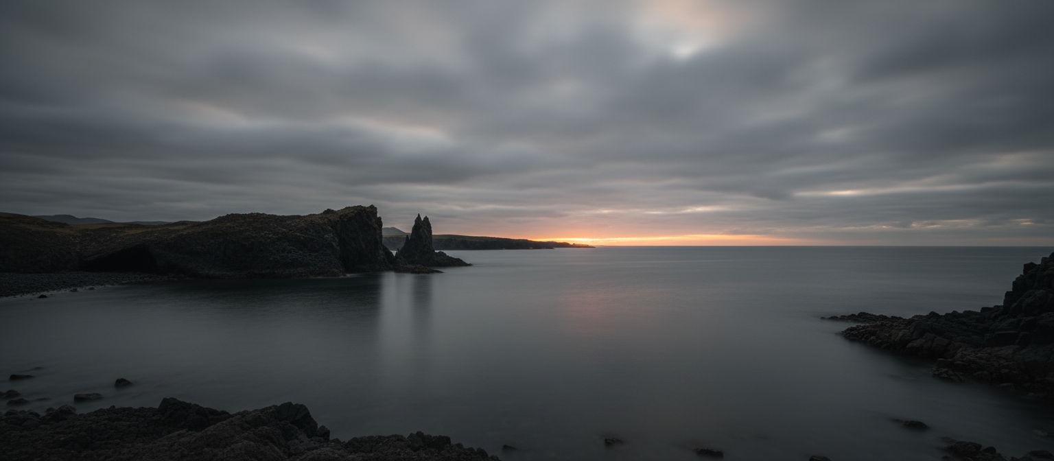 Dark rocky coastline at dawn with a thin band of warm light on the horizon