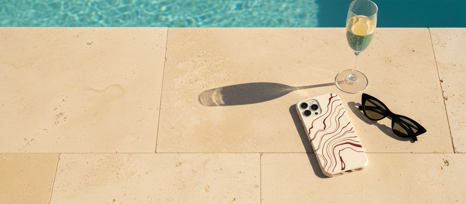 Sunlit travertine pool terrace with an iPhone in a cream sotto voce case with oxblood ink lines, a flute of champagne and black cat-eye sunglasses