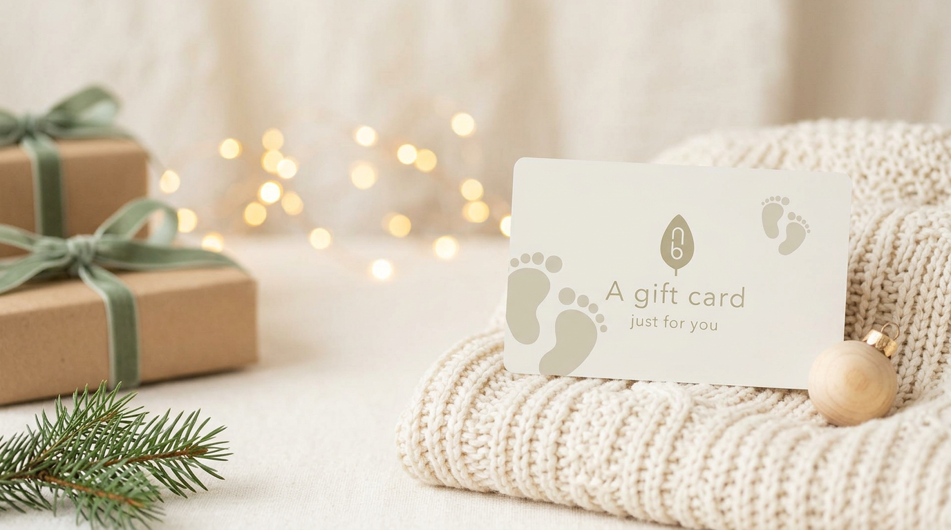 Natural Baby Shower gift card propped on a cream knitted blanket beside a wooden bauble, with kraft-wrapped presents and fairy lights behind