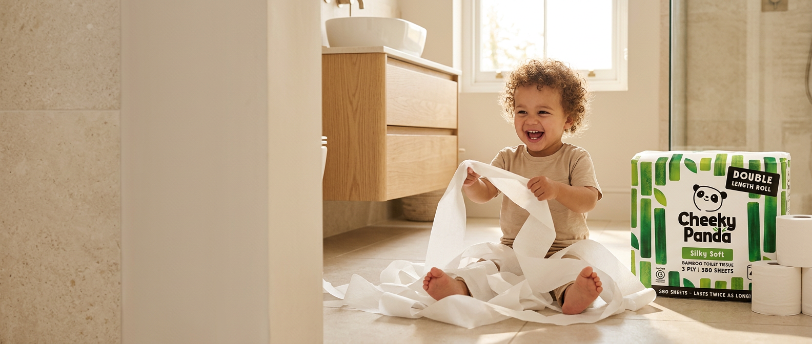 A laughing toddler playing with unrolled Cheeky Panda toilet paper beside a Double-Length Roll pack